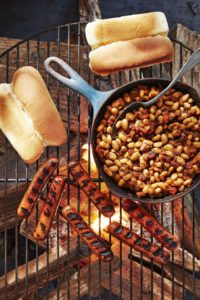 Beans in a cast iron pan over a campfire with hot dogs and buns next to it